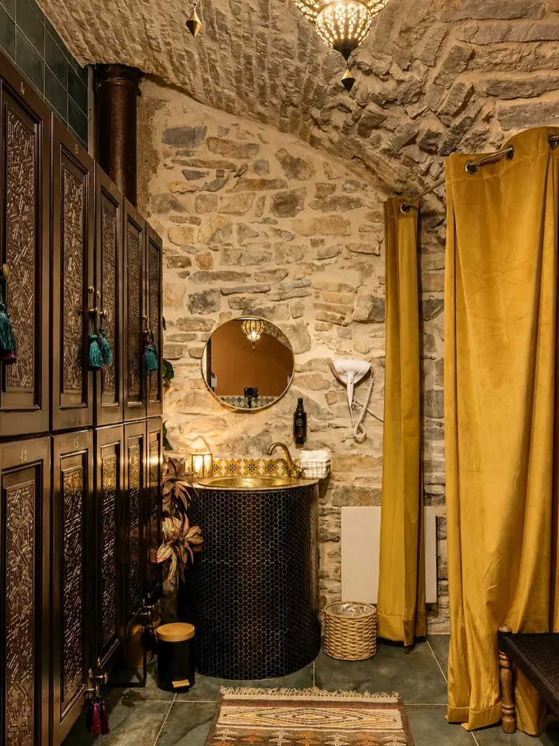 An elegant changing room with vaulted stone walls, carved wooden lockers, a round basin, and mustard-yellow curtains.