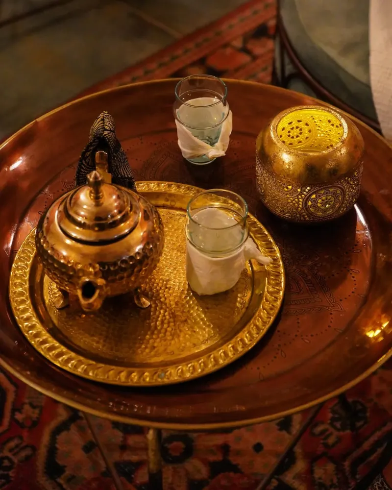 A traditional tea set on a golden brass tray, featuring a teapot, glasses, and a lantern in a warm setting.